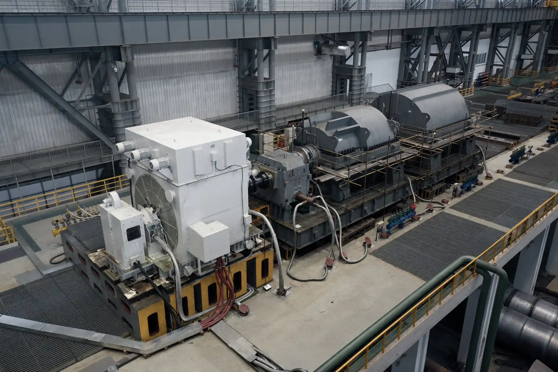 Overhead view of a motor test bay: a large electric motor coupled through a gearbox to two machine casings on a foundation, with hoses and cable trays on the floor.