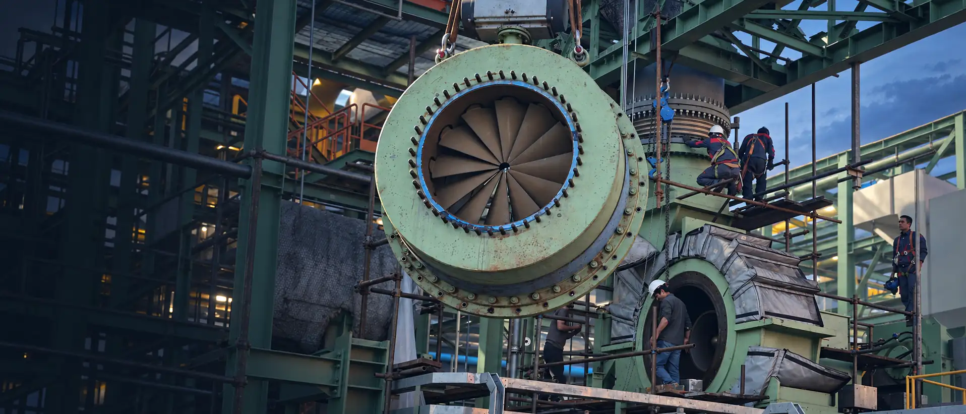 Large green machine casing lifted by crane beside an open casing on a steel structure, with technicians in hard hats and harnesses on the scaffolding around it at dusk.