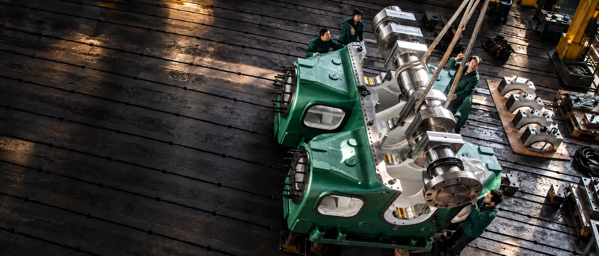 Overhead view of an assembly workshop: a polished crankshaft suspended from crane slings over the open green frame of a large reciprocating machine, with technicians in green workwear beside it and bearing caps laid out on the floor.